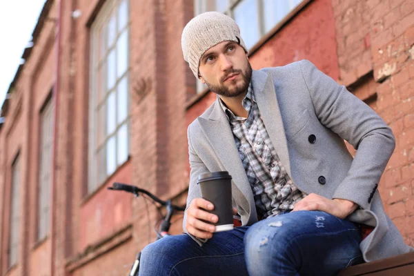 Bello giovane in cappotto grigio e cappello seduto su una panchina rilassato bere caffè e pensare vicino alla sua bicicletta . — Foto Stock