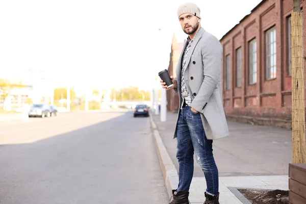 Schöner junger Mann in grauem Mantel und Hut überquert mit einer Tasse Kaffee die Straße. — Stockfoto