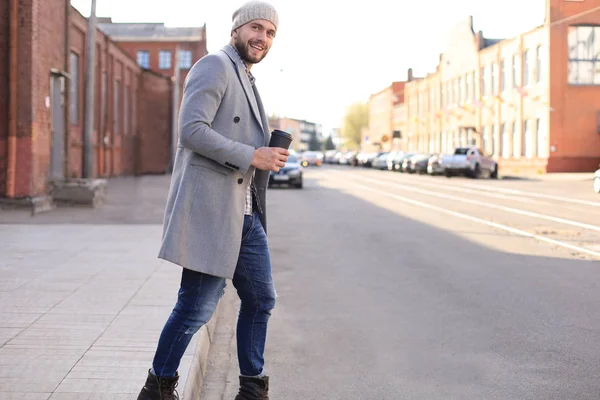 Schöner junger Mann in grauem Mantel und Hut überquert mit einer Tasse Kaffee die Straße. — Stockfoto