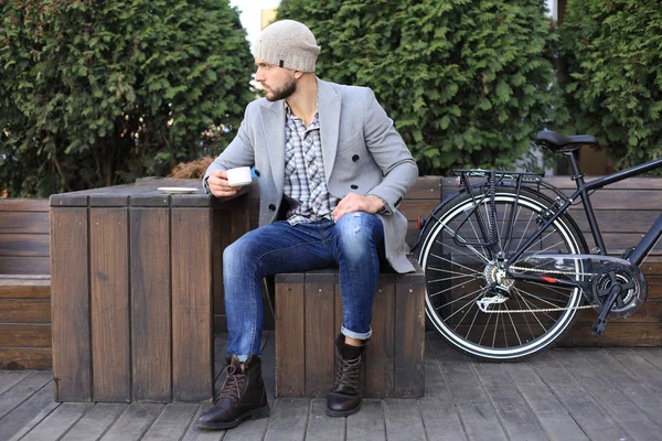 Schöner junger Mann in grauem Mantel und Hut sitzt entspannt auf einer Bank, trinkt Kaffee und denkt in der Nähe seines Fahrrads. — Stockfoto