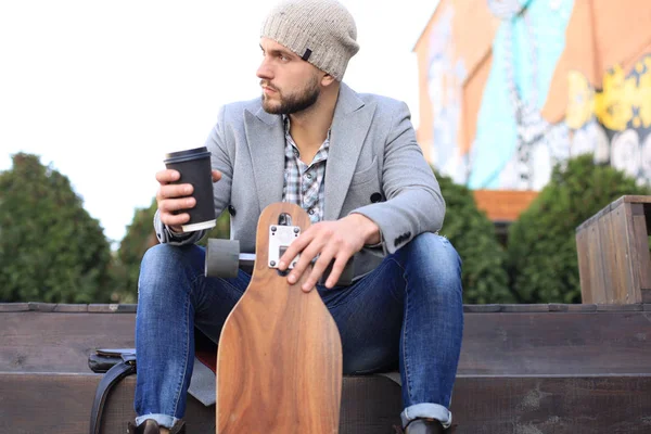 Bello giovane in cappotto grigio e cappello, a riposo, seduto con longboard bere caffè. Concetto di skateboard urbano . — Foto Stock