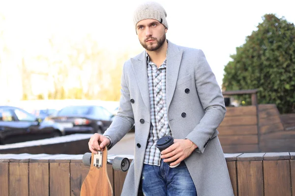 Schöner junger Mann in grauem Mantel und Hut, der Kaffee trinkt, sich ausruht und mit Longboard steht. städtisches Skateboarding-Konzept. — Stockfoto