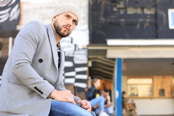 Gutaussehender junger Mann in grauem Mantel und Hut, auf einer Bank sitzend. Stadtentwicklungskonzept. — Stockfoto