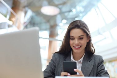 Ofiste bilgisayarın yanında akıllı telefon kullanan gülümseyen genç bir iş kadını..