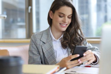 Ofiste bilgisayarın yanında akıllı telefon kullanan gülümseyen genç bir iş kadını..