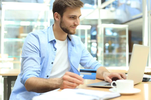 Hombre sonriente sentado en la oficina y paga con tarjeta de crédito con su computadora portátil. — Foto de Stock