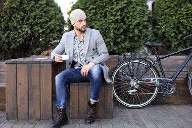 Gri ceketli ve şapkalı yakışıklı genç adam bir bankta oturarak rahat bir kahve içiyor ve bisikletinin yanında düşünüyor..