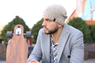 Gri ceketli ve şapkalı yakışıklı genç adam, dinleniyor, uzun tahtayla oturuyor. Kentsel kaykay konsepti.