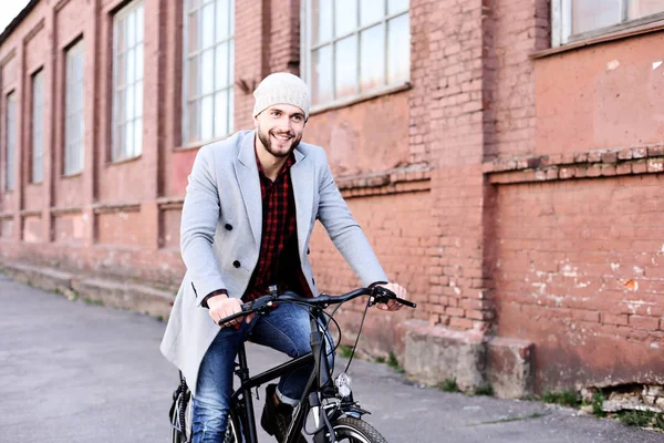 灰色のコートと帽子をかぶったハンサムな若者が市内の自転車通りに乗る. — ストック写真