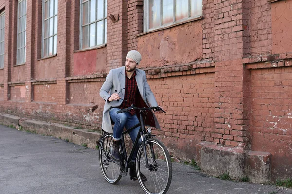 Красивий молодий чоловік у сірому пальто і капелюсі їде на велосипедній вулиці в місті . — стокове фото
