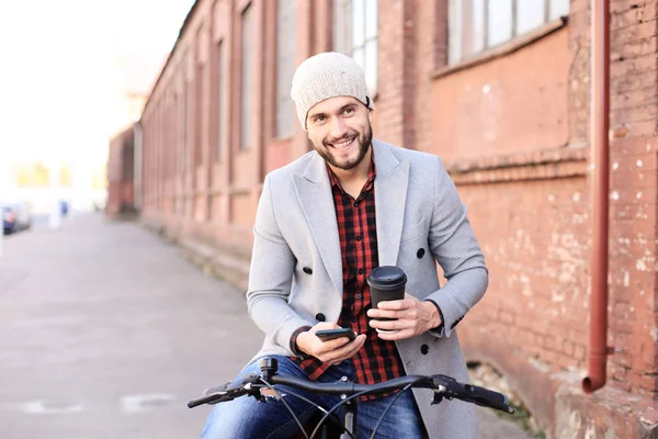 灰色のコートと帽子を着て、彼の横に彼の自転車を押しながら電話を見てハンサムな若者. — ストック写真