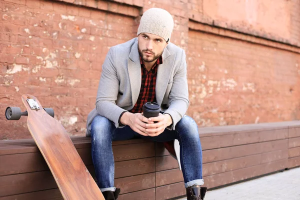 Beau jeune homme en manteau et chapeau gris, au repos, assis avec longboard. Concept de skateboard urbain . — Photo