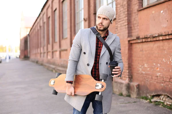 Bello giovanotto in cappotto grigio e cappello che cammina per strada, usando il longboard . — Foto Stock