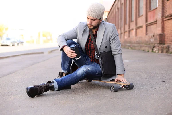 Schöner junger Mann in grauem Mantel und Hut, der auf dem Longboard auf der Straße in der Stadt sitzt. städtisches Skateboarding-Konzept. — Stockfoto