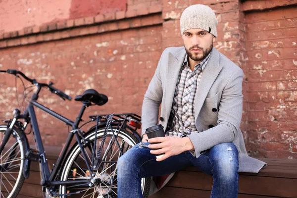 Schöner junger Mann in grauem Mantel und Hut sitzt entspannt auf einer Bank, trinkt Kaffee und denkt in der Nähe seines Fahrrads. — Stockfoto