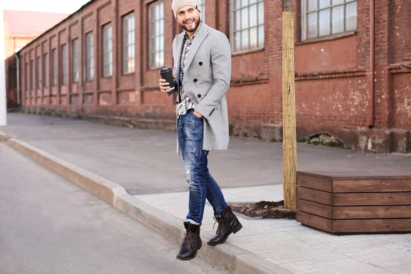 Beau jeune homme en manteau gris et chapeau traversant la rue avec une tasse de café . — Photo