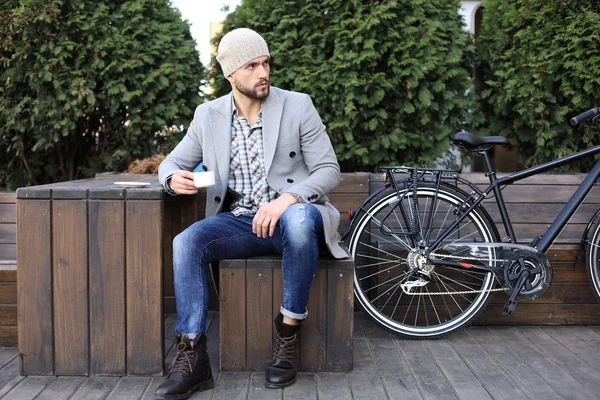 Schöner junger Mann in grauem Mantel und Hut sitzt entspannt auf einer Bank, trinkt Kaffee und denkt in der Nähe seines Fahrrads. — Stockfoto