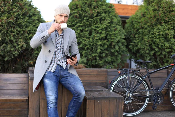 Schöner junger Mann in grauem Mantel und Hut mit Smartphone, ruht sich aus und lächelt, während er im Freien neben seinem Fahrrad sitzt. — Stockfoto