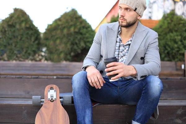 Bello giovane in cappotto grigio e cappello, a riposo, seduto con longboard bere caffè. Concetto di skateboard urbano . — Foto Stock