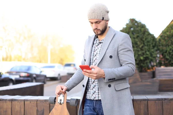 Gutaussehender junger Mann in grauem Mantel und Hut mit Smartphone, liegend, stehend mit Longboard. städtisches Skateboarding-Konzept. — Stockfoto