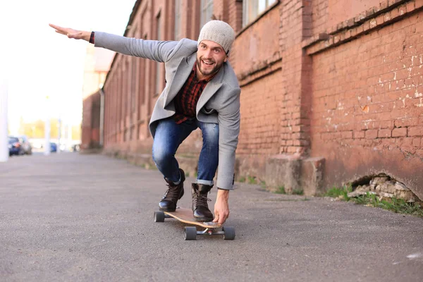 街の路上で長いボードに灰色のコートと帽子をかぶったハンサムな若者。アーバンスケートボードコンセプト. — ストック写真