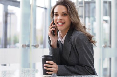 Ofiste bilgisayarın yanında akıllı telefon kullanan gülümseyen genç bir iş kadını..