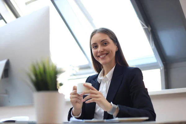 Atraktivní usměvavá žena, která sedí v kanceláři s šálkem kávy. — Stock fotografie