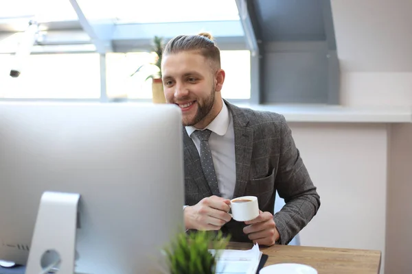 Joven hombre de negocios beber café en la oficina mientras escribe en el ordenador . — Foto de Stock