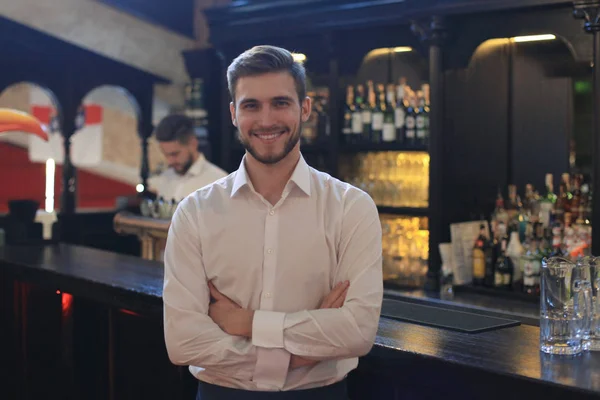 Successful small business owner standing with crossed arms with employee in background preparing coffee.