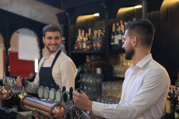 Bello barista sorride e riempie un bicchiere di birra mentre in piedi al bancone del bar in pub . — Foto Stock