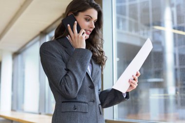 Ofiste bilgisayarın yanında akıllı telefon kullanan gülümseyen genç bir iş kadını..