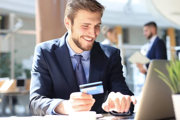 Hombre sonriente sentado en la oficina y paga con tarjeta de crédito con su computadora portátil. — Foto de Stock