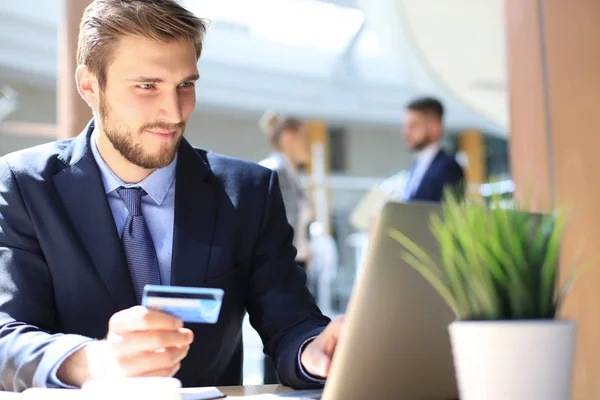 Hombre sonriente sentado en la oficina y paga con tarjeta de crédito con su computadora portátil. — Foto de Stock
