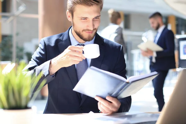 Молодий бізнесмен п'є каву в офісі, перевіряючи блокнот . — стокове фото