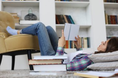 Evde okuyan, üniversite sınavlarına hazırlanan, rahat iç mekana karşı yerde yatan, kitap yığınıyla çevrili genç kız öğrenci.