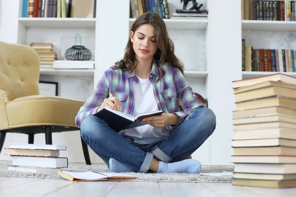 Young female student studying at home, writing article for publication, sitting on floor against cozy domestic interior, surrounded with pile of books. — Stock Photo, Image