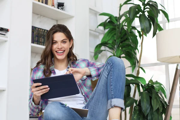 Sorrindo jovem mulher sentada no chão na sala de estar, estudante estudando em casa trabalhando com seu tablet . — Fotografia de Stock