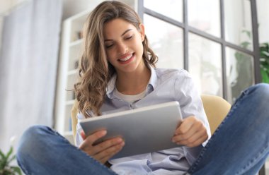 Gülümseyen genç kadın oturma odasında koltukta oturan ve onun tablet ile kullanarak.