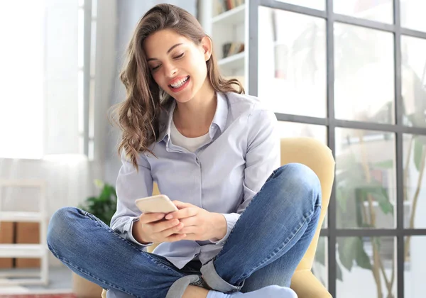 Sorridente giovane donna seduta sulla poltrona in soggiorno e utilizzando con il suo telefono cellulare . — Foto Stock