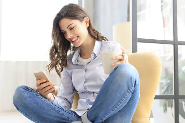 Mujer joven sonriente sentada en el sillón en la sala de estar y usando con su teléfono móvil . —  Fotos de Stock