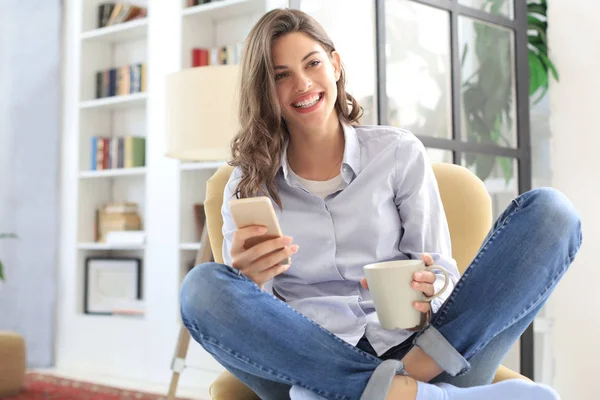 Усміхнена молода жінка сидить у кріслі у вітальні і використовує зі своїм мобільним телефоном . — стокове фото