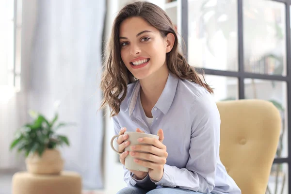 Mujer joven sonriente sentada en el sillón de la sala de estar, sosteniendo una taza de café y mirando a la cámara . —  Fotos de Stock