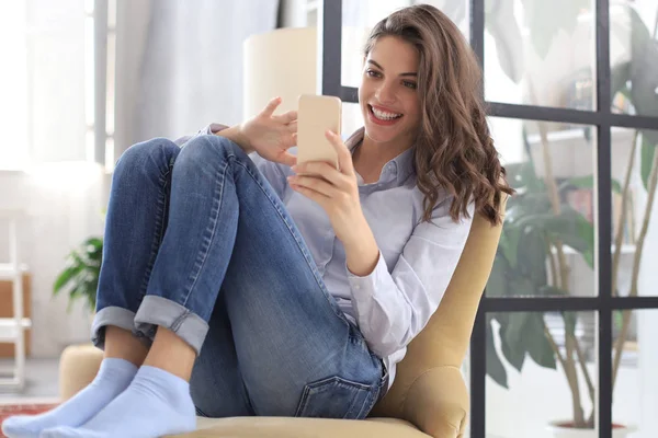 Lächelnde junge Frau sitzt im Sessel im Wohnzimmer und benutzt mit ihrem Handy. — Stockfoto