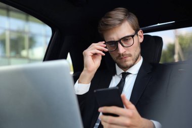 Arabada otururken ve dizüstü bilgisayar kullanırken onun akıllı telefon bakarak tam takım elbise yakışıklı kendinden emin adam.