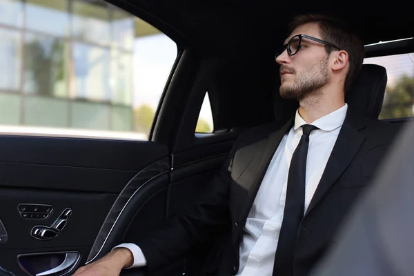 Pensativo empresário confiante em terno completo com óculos olhando para longe enquanto sentado no carro . — Fotografia de Stock