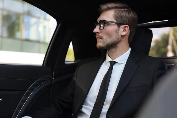 Homme d'affaires réfléchi et confiant en costume complet avec des lunettes regardant loin tout en étant assis dans la voiture . — Photo