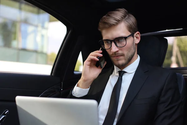 Bello uomo d'affari fiducioso in giacca e cravatta che parla su smartphone e lavora con il computer portatile mentre è seduto in macchina . — Foto Stock
