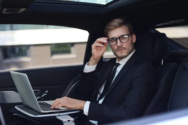 Doordachte jonge zakenman die hand op bril houdt terwijl hij in de Lux-auto zit en zijn laptop gebruikt. — Stockfoto