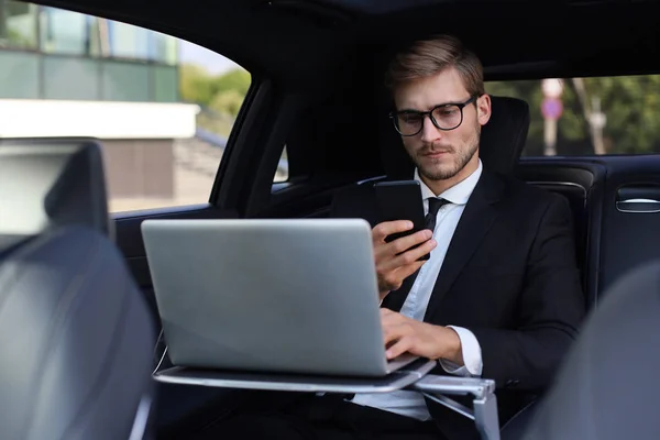 Knappe Zelfverzekerde man in volle pak kijken naar zijn slimme telefoon terwijl zitten in de auto en het gebruik van laptop. — Stockfoto