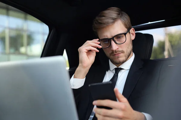 Snygg självsäker man i full kostym tittar på hans smarta telefon när du sitter i bilen och använda laptop. — Stockfoto